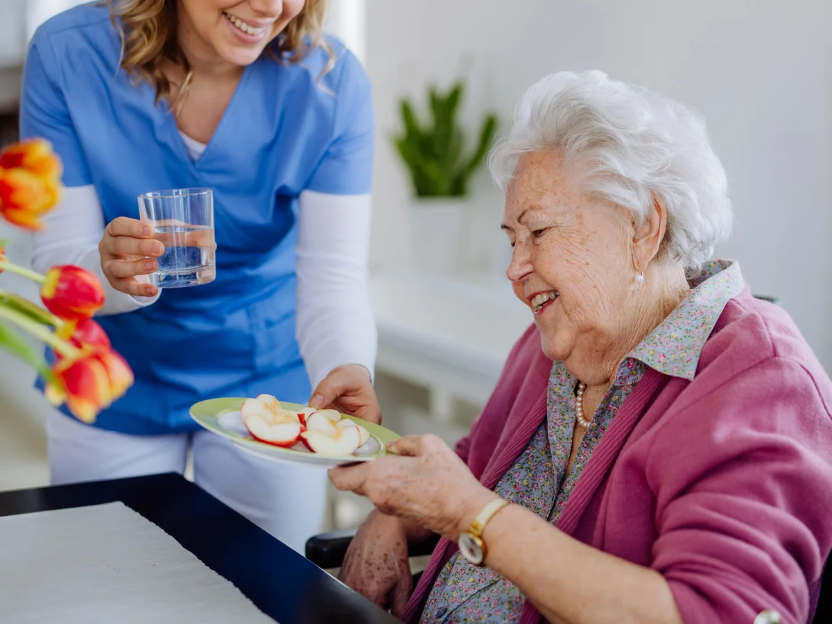 Pflegekraft reicht Seniorin Apfelscheiben und Wasser